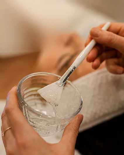 Mão segurando tigela de vidro com o peeling e o pincel, cliente deitada ao fundo